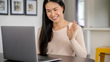 A patient sits at her kitchen table with a laptop out in front of her. She is dressed casually and waiving "goodbye" during a remote appointment with her doctor via a video call.