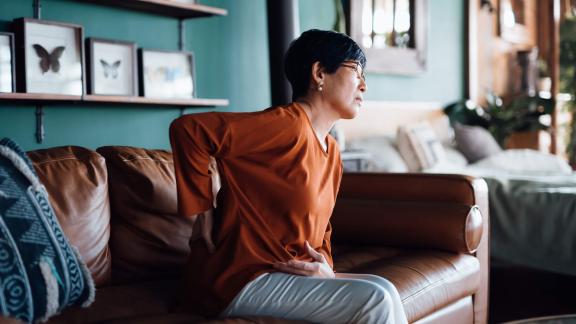 Woman sitting on sofa rubbing her back, looking unwell.