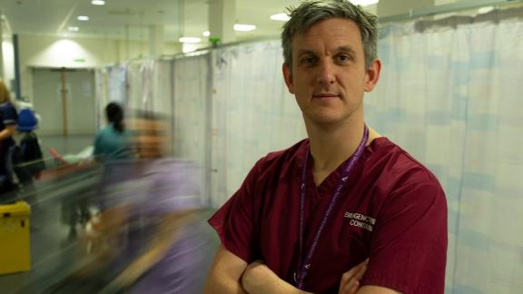 A photo of Dr Chris Pickering, wearing medical scrubs, photographed in a busy hospital environment.