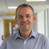 Accessibility description: A photo of Dr Mark Simmonds, a middle-aged man with short dark hair, smiling and looking towards the camera while wearing a checked light blue shirt, with a hospital interior in the background.